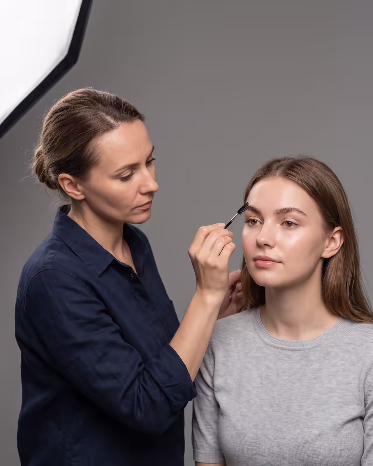 A female makeup artist wearing a navy blue shirt grooms a young woman's eyebrows using an eyebrow spoolie brush in a photo studio. The model sits facing forward in a grey crewneck top against a neutral background beside a large softbox light.