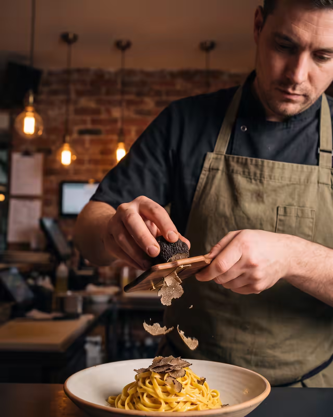 An adult male chef wearing a black short-sleeved shirt and an olive green apron shaves fresh black truffle over a plated swirl of yellow pasta in a ceramic bowl using a wooden truffle slicer. Slices of truffle fall onto the pasta dish within a warm restaurant interior.