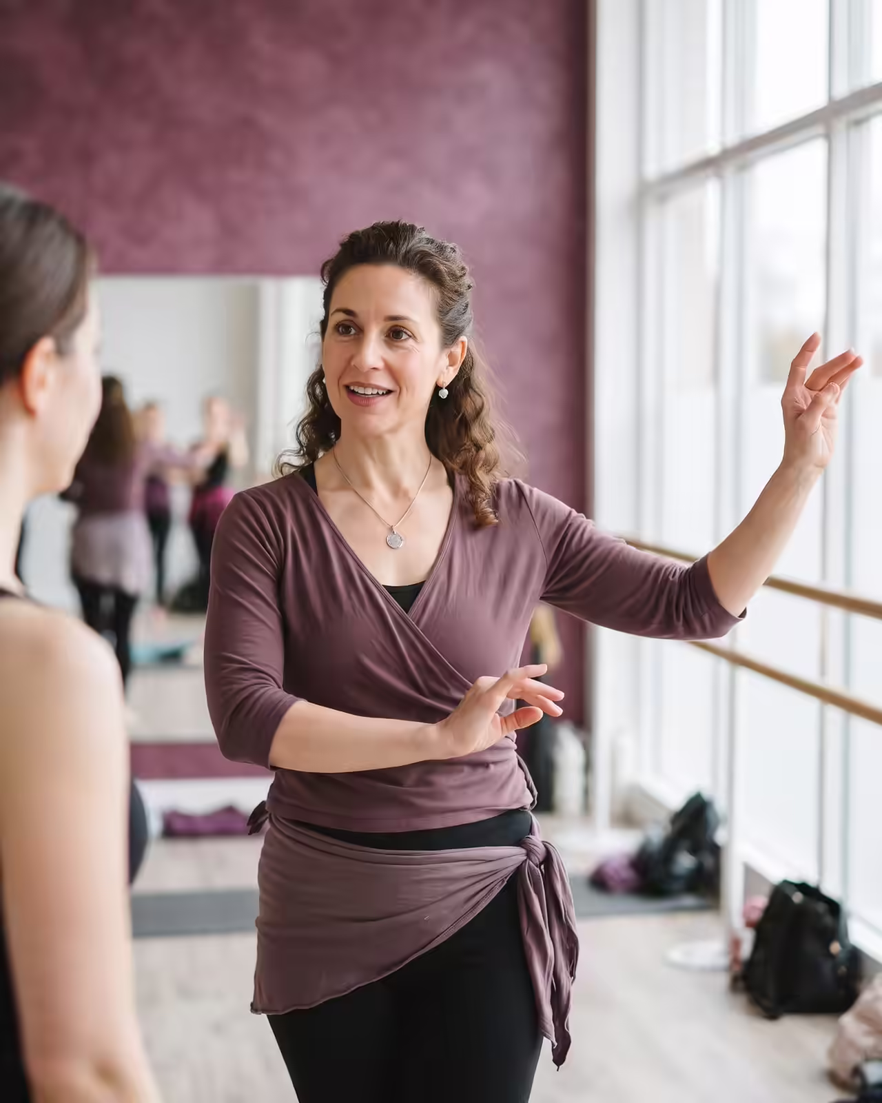 A smiling dance teacher with curly brown hair wearing a mauve wrap top, matching hip scarf, and black leggings demonstrates an expressive hand gesture while speaking with a student in a sunlit dance studio with mirrors and a ballet barre.