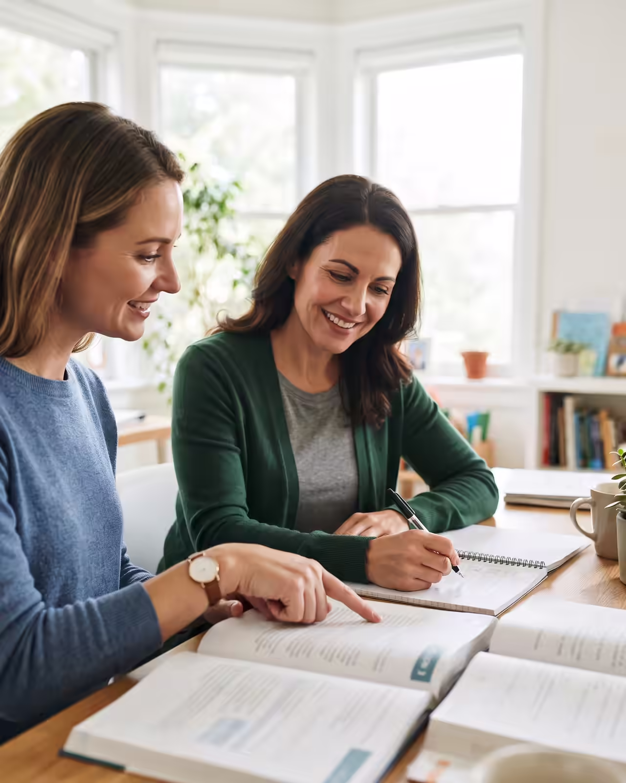 A smiling female tutor in a blue sweater points to a passage in an open textbook while guiding an adult female student in a green cardigan who takes notes with a pen in a spiral notebook at a sunlit wooden table.